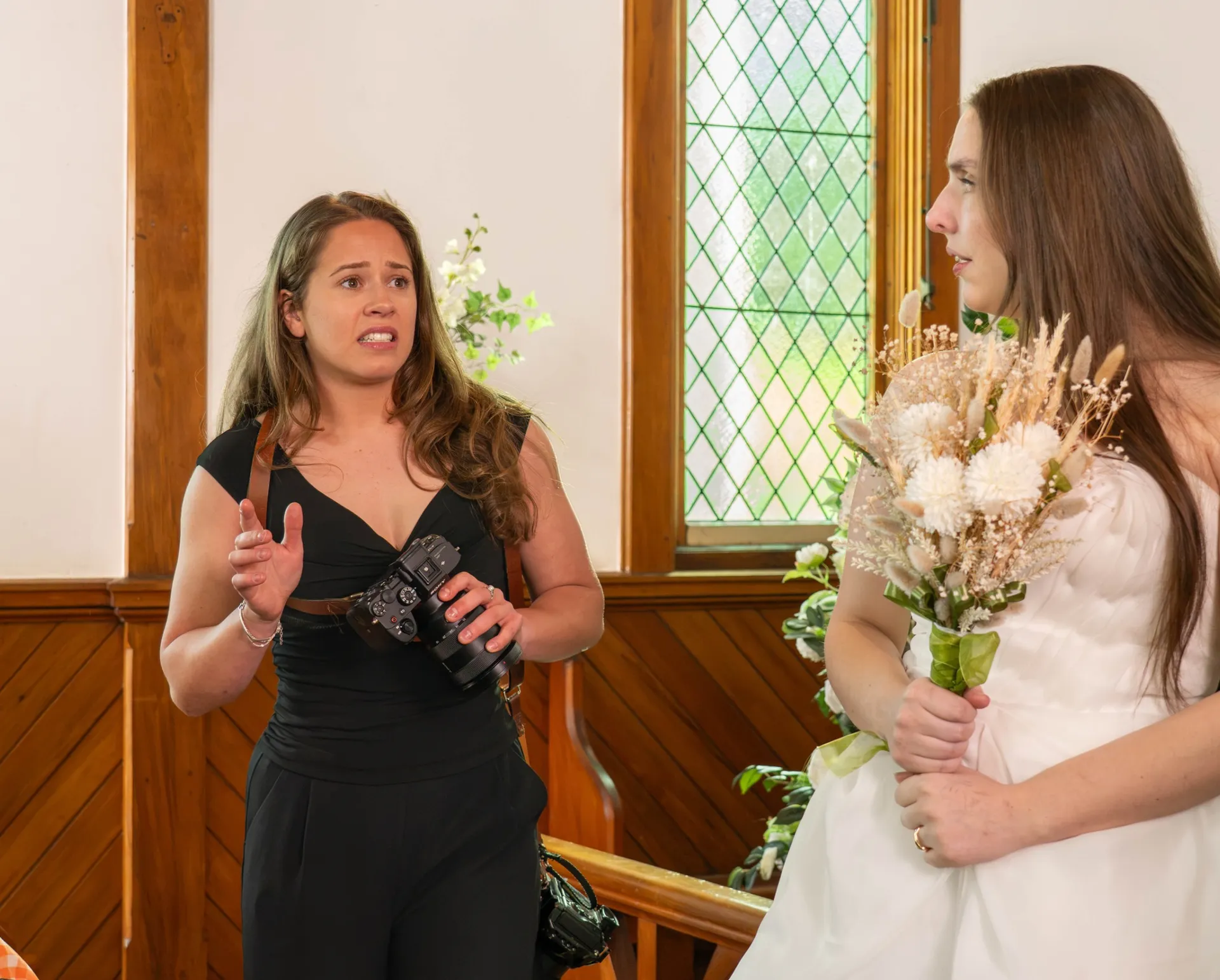 A wedding photographer holding her camera talks with the bride in a small chapel, guests seated beside them